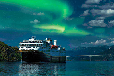 Havila Kystruten Schiff fährt unter leuchtenden grünen Nordlichtern (Aurora Borealis) und einem dramatischen Himmel entlang der norwegischen Küste.