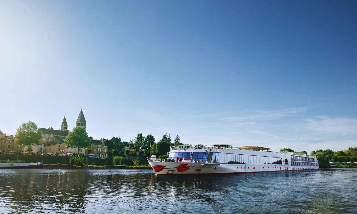 Weißes A-ROSA Flusskreuzfahrtschiff mit rotem Kussmund fährt auf einem Fluss an einer Stadt mit Kirchttürmen unter blauem Himmel vorbei.