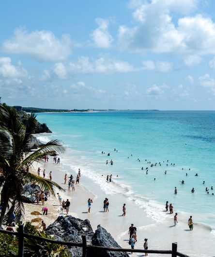 Ein belebter, weißer Sandstrand mit türkisfarbenem Meer. Viele Menschen schwimmen im Wasser oder spazieren am Ufer, eingerahmt von Felsen und Palmen.