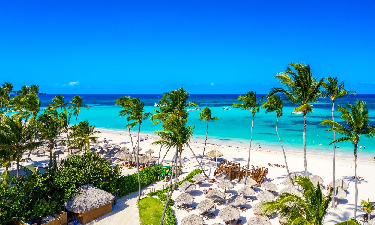 Tropischer Sandstrand aus der Vogelperspektive mit vielen Palmen, Reetdach-Schirmen und türkisblauem Meer unter strahlend blauem Himmel.