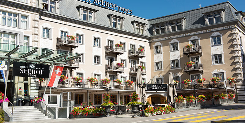Das Hotel Seehof Davos in der Schweiz, ein klassisches Hotelgebäude mit mehreren Etagen, zahlreichen Balkonen und vielen Blumenkästen voller bunter Blüten. An der Vorderseite sind die Hotelrestaurants mit Außensitzplätzen und Sonnenschirmen zu erkennen. Eine Schweizer Flagge weht links vor dem Eingang, und der Himmel ist klar blau.